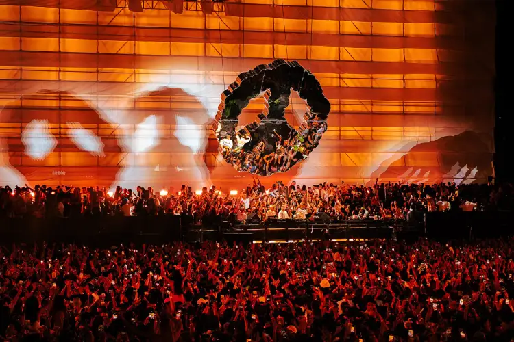 Large crowd with raised hands watching a concert stage with massive orange lighting and a circular design element suspended above Large crowd with raised hands watching a concert stage with massive orange lighting and a circular design element suspended above