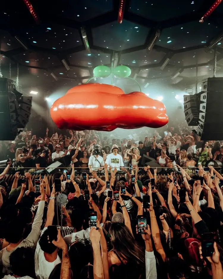 Large crowd with raised hands at an indoor concert venue beneath an illuminated red balloon installation and green decorative elements Large crowd with raised hands at an indoor concert venue beneath an illuminated red balloon installation and green decorative elements