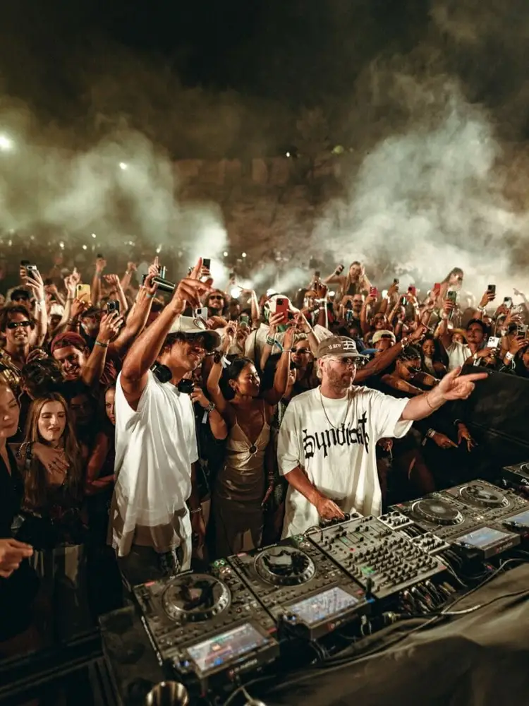 DJs perform at a nighttime outdoor music festival as a large crowd with raised arms and phones surrounds the stage in clouds of powder or smoke DJs perform at a nighttime outdoor music festival as a large crowd with raised arms and phones surrounds the stage in clouds of powder or smoke