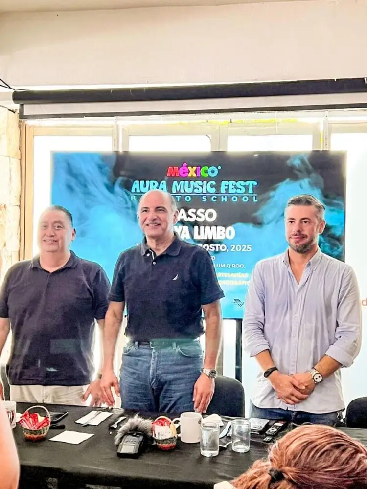 Three men stand at a table in front of a screen displaying a poster for Mexico Aura Music Fest event Three men stand at a table in front of a screen displaying a poster for Mexico Aura Music Fest event