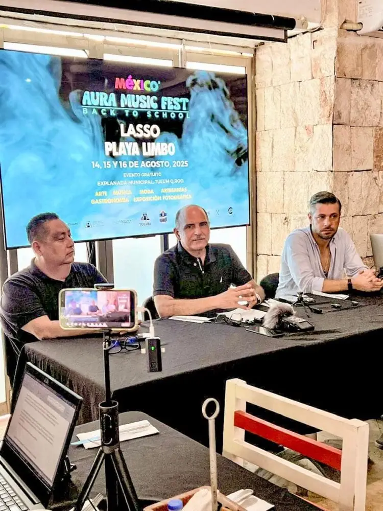 Three men sit at a table during a press conference with a screen behind them displaying information about a music festival Three men sit at a table during a press conference with a screen behind them displaying information about a music festival