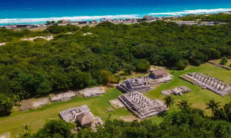 Aerial view of ancient stone ruins scattered across a grassy field surrounded by dense green jungle, with a coastal town and turquoise ocean visible in the dist