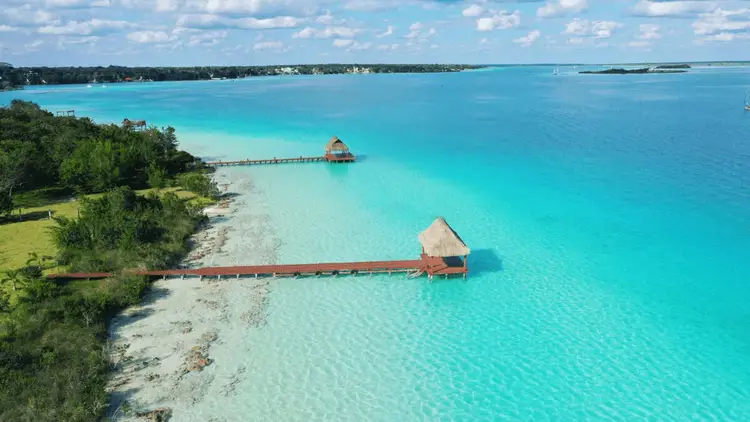 Thatched-roof bungalows extend on wooden piers into turquoise shallow water beside a forested shoreline and sandy beach