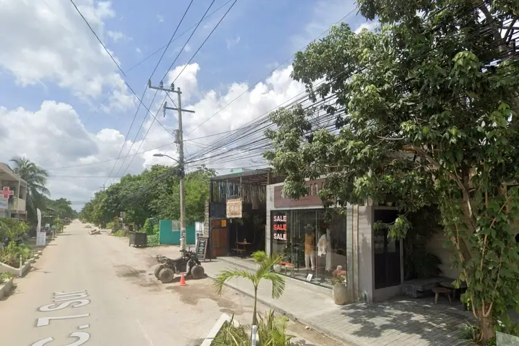 Storefronts line a sandy street with power lines overhead and vegetation on the right side under a partly cloudy sky