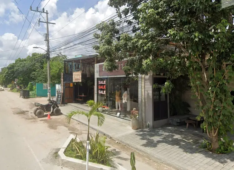 Small shops and businesses line a residential street with overgrown vines covering structures, power lines overhead, and a motorcycle parked on the unpaved road