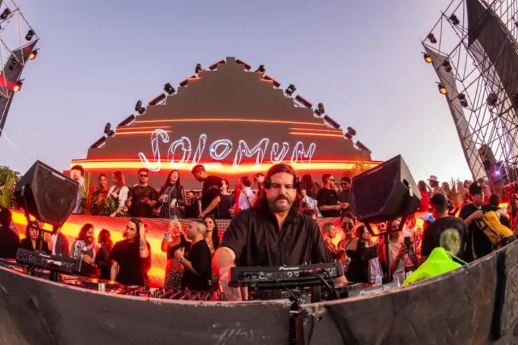 DJ performing at an outdoor music festival stage with neon signage and crowds gathered under evening lights DJ performing at an outdoor music festival stage with neon signage and crowds gathered under evening lights
