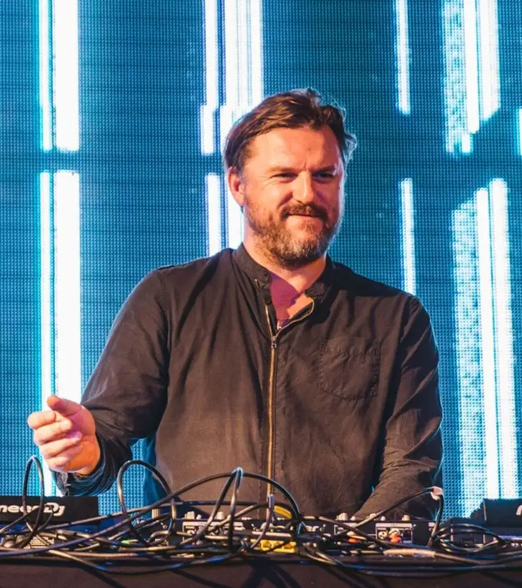 Man in black jacket gesturing while standing behind DJ equipment in front of a blue LED screen display Man in black jacket gesturing while standing behind DJ equipment in front of a blue LED screen display