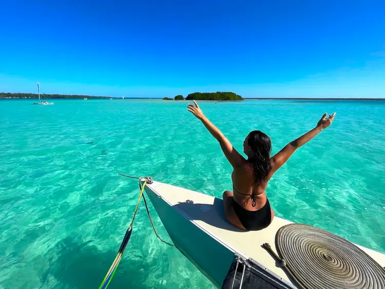 Person with arms raised sits on a boat in clear turquoise water with a forested island and sailboats visible in the distance