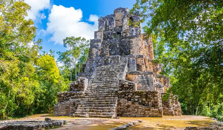 Ancient stone pyramid with steep stairs rises from a cleared plaza surrounded by dense jungle vegetation under blue sky with white clouds