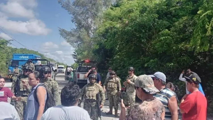 Armed personnel in military fatigues stand on a rural road alongside civilians and vehicles during an apparent eviction operation
