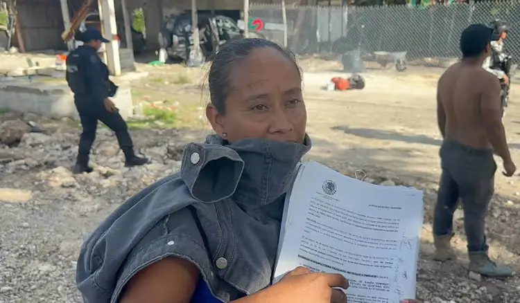 Woman holds an official document while standing in a sandy lot with other people and structures visible in the background