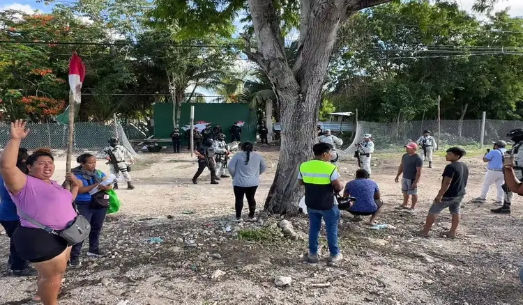 Group of people gathered in a dusty outdoor area beneath a large tree, with green fencing and structures visible in the background