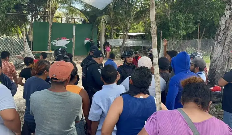 Police and residents gather in a dusty outdoor area surrounded by palm trees during what appears to be an eviction or displacement situation