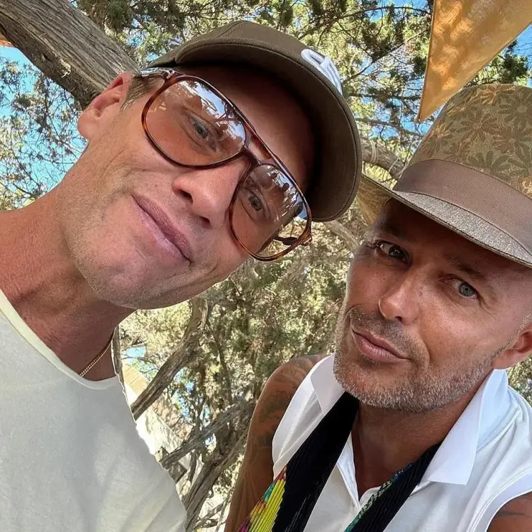 Two men wearing hats and glasses take a selfie outdoors under tree branches Two men wearing hats and glasses take a selfie outdoors under tree branches
