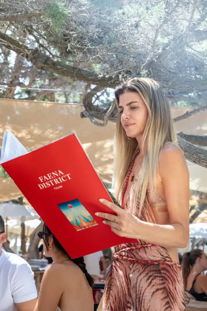 Woman holding a red book titled "Faena District" outdoors under a tree at what appears to be an event or gathering Woman holding a red book titled "Faena District" outdoors under a tree at what appears to be an event or gathering