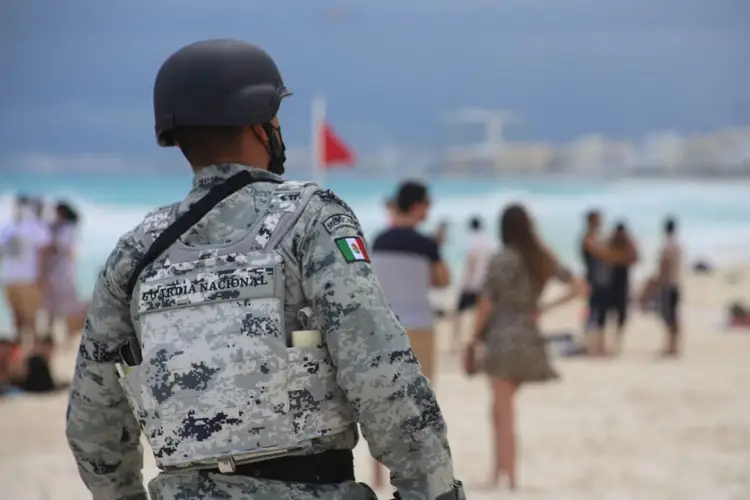 Military personnel in camouflage uniform stands on a beach watching beachgoers, with a flag visible in the background Military personnel in camouflage uniform stands on a beach watching beachgoers, with a flag visible in the background