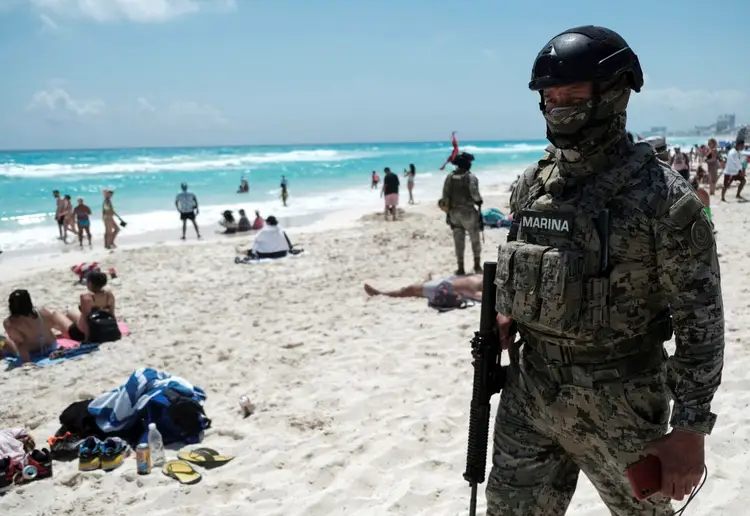 Armed military personnel in tactical gear stands on a sandy beach among beachgoers and swimmers in the turquoise water Armed military personnel in tactical gear stands on a sandy beach among beachgoers and swimmers in the turquoise water