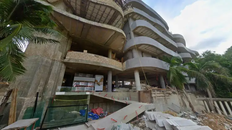 Abandoned multi-story concrete building with curved balconies, construction debris, and palm trees visible on the overgrown hillside