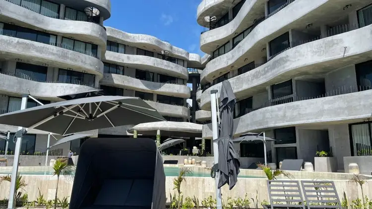Curved concrete apartment buildings with multiple balconies overlook a courtyard with umbrellas and lounge chairs below