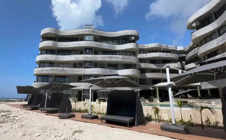 Modern curved apartment building with white balconies overlooking a beach, with dark umbrellas and seating structures in the sand below