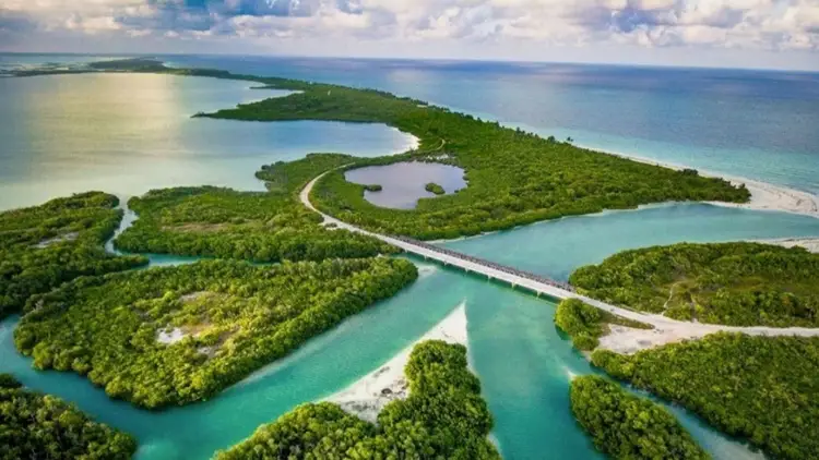 Aerial view of a tropical island with dense green vegetation, turquoise waterways, white sand beaches, and a winding road cutting through mangrove forests