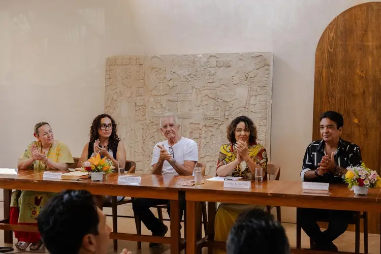 Five people sit at a long table during a panel discussion, with audience members visible in the foreground and an ancient stone carving mounted on the wall behi Five people sit at a long table during a panel discussion, with audience members visible in the foreground and an ancient stone carving mounted on the wall behi
