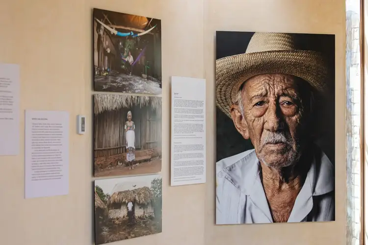 Gallery wall displays portrait photographs and text panels, including a large close-up of an elderly man wearing a straw hat and light-colored shirt Gallery wall displays portrait photographs and text panels, including a large close-up of an elderly man wearing a straw hat and light-colored shirt