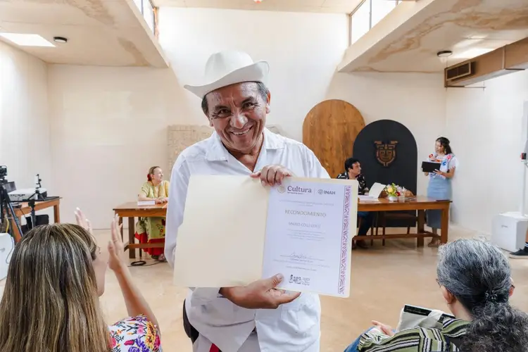 Man in white hat and shirt displays a certificate to two people seated in front of him during an indoor ceremony Man in white hat and shirt displays a certificate to two people seated in front of him during an indoor ceremony