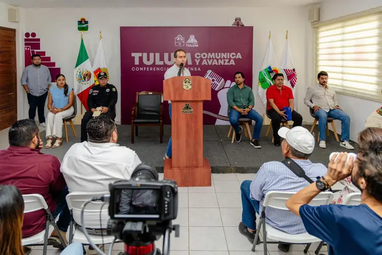 Man in orange shirt speaks at wooden podium during press conference with seated officials and audience members, Mexican and white flags visible behind him Man in orange shirt speaks at wooden podium during press conference with seated officials and audience members, Mexican and white flags visible behind him