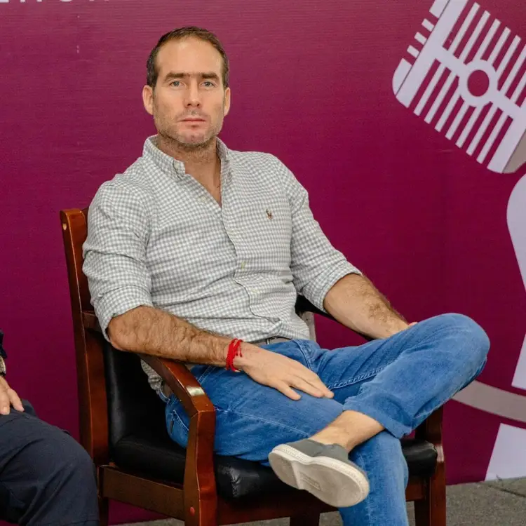 Man in checkered shirt and jeans sitting in a chair against a magenta background with partial graphic visible on the right Man in checkered shirt and jeans sitting in a chair against a magenta background with partial graphic visible on the right
