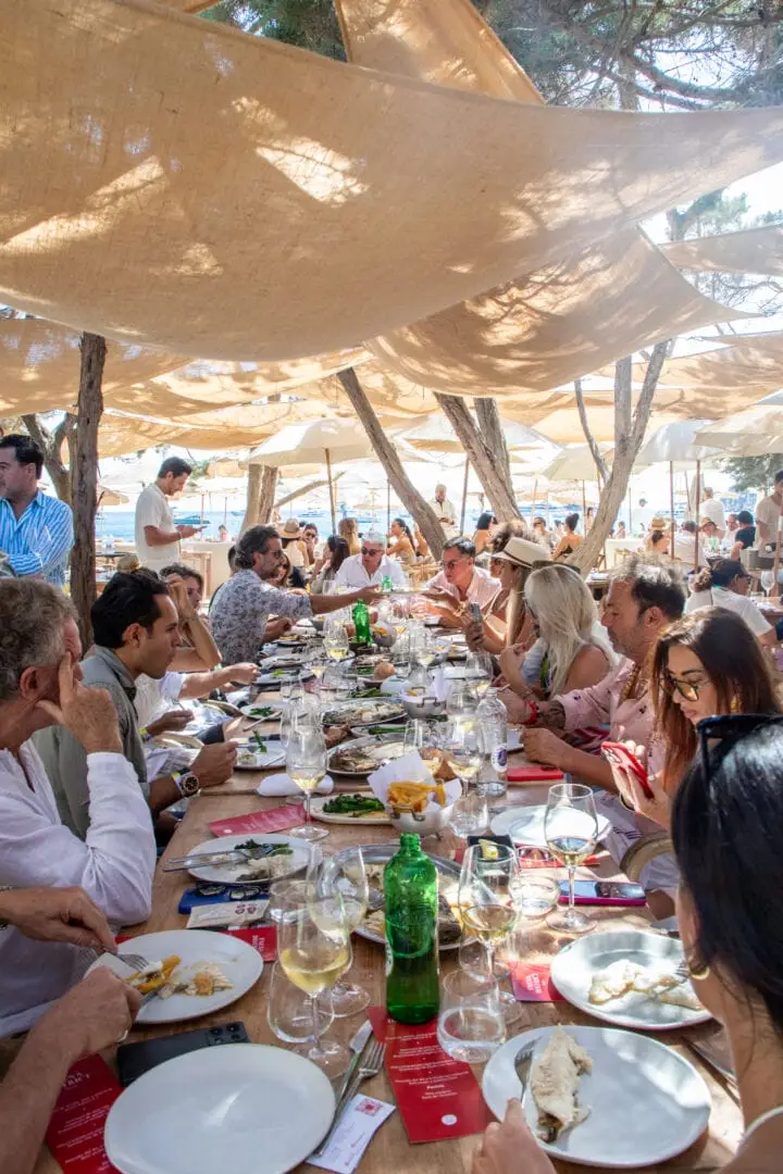 Large group of people dining under canvas canopies at a beachfront restaurant with water visible in the background Large group of people dining under canvas canopies at a beachfront restaurant with water visible in the background