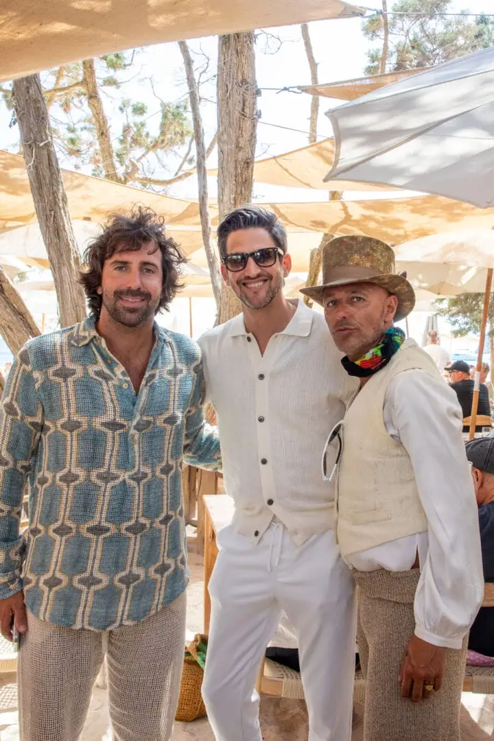 Three men stand together under tan shade sails at an outdoor event with trees and other guests visible in the background Three men stand together under tan shade sails at an outdoor event with trees and other guests visible in the background