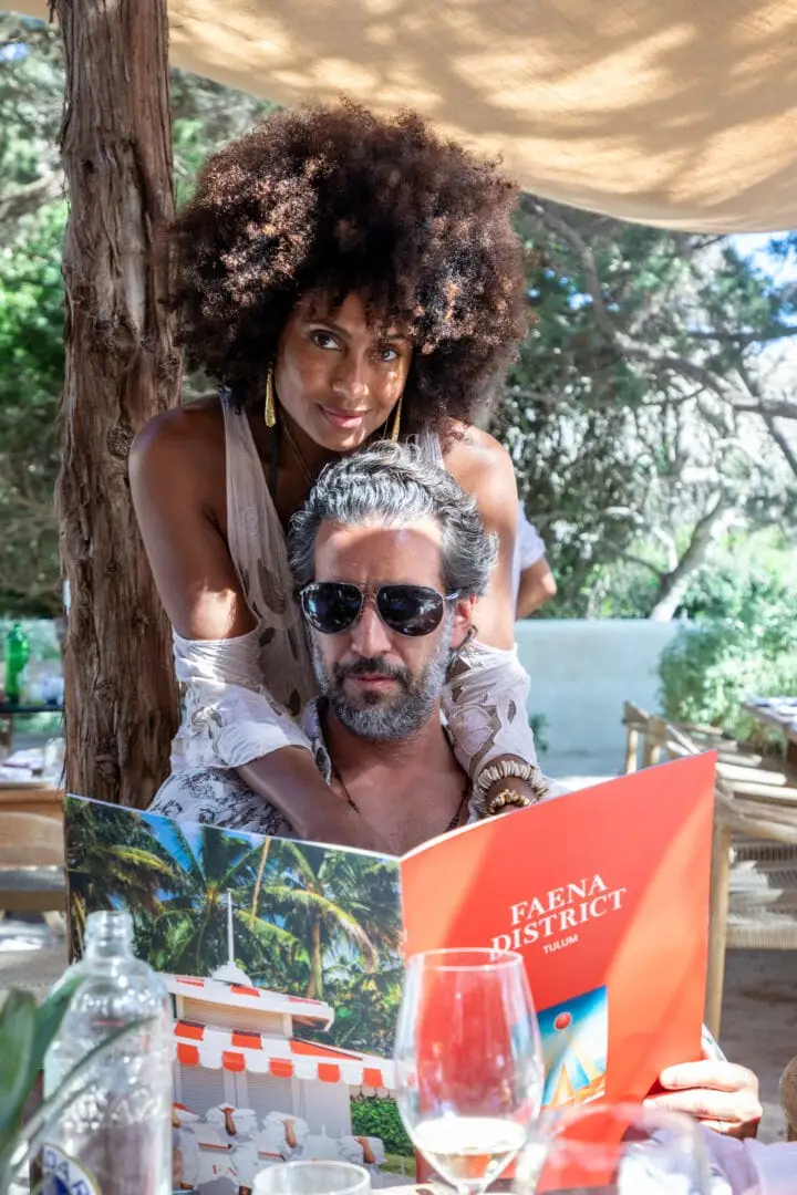 Two people posing together outdoors under a wooden structure, one holding an orange Faena District publication, with a shaded patio and trees visible in the bac Two people posing together outdoors under a wooden structure, one holding an orange Faena District publication, with a shaded patio and trees visible in the bac