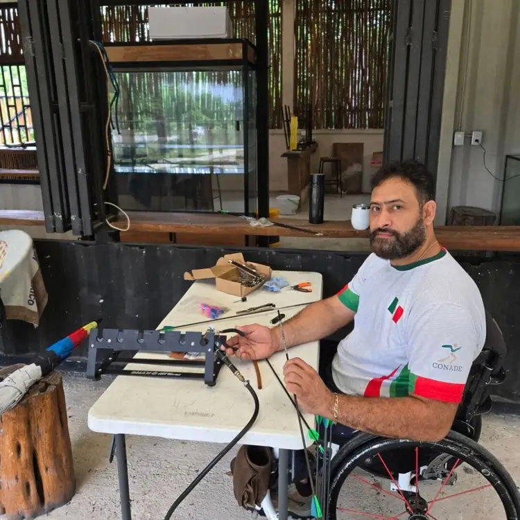 Man in a wheelchair works with archery equipment at a table in an indoor facility with an aquarium and decorative screens visible in the background Man in a wheelchair works with archery equipment at a table in an indoor facility with an aquarium and decorative screens visible in the background
