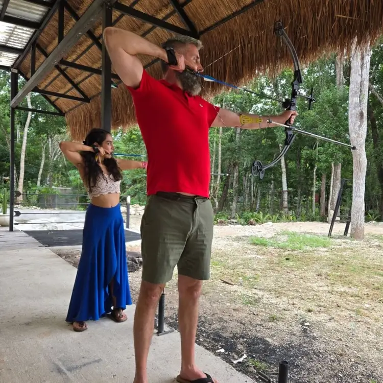 Man in red shirt draws a compound bow while a woman watches under a thatched shelter in a wooded area Man in red shirt draws a compound bow while a woman watches under a thatched shelter in a wooded area