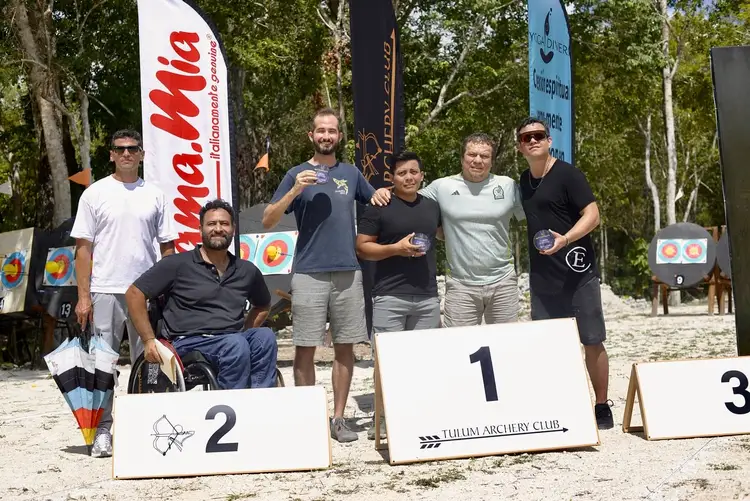 Six people stand on a podium at an outdoor archery competition, with branded banners and trees in the background Six people stand on a podium at an outdoor archery competition, with branded banners and trees in the background