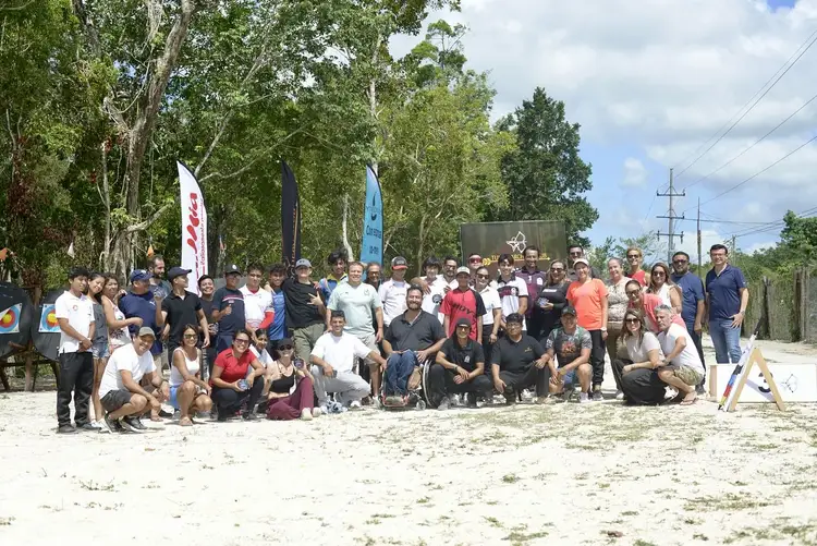 Large group of people gathered on a sandy beach with trees and power lines in the background, some standing and others crouching or sitting in the sand Large group of people gathered on a sandy beach with trees and power lines in the background, some standing and others crouching or sitting in the sand