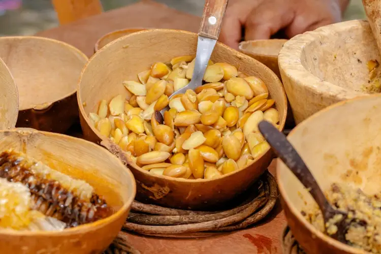 A person scoops roasted cashews from a wooden bowl surrounded by other bowls of nuts and seeds A person scoops roasted cashews from a wooden bowl surrounded by other bowls of nuts and seeds