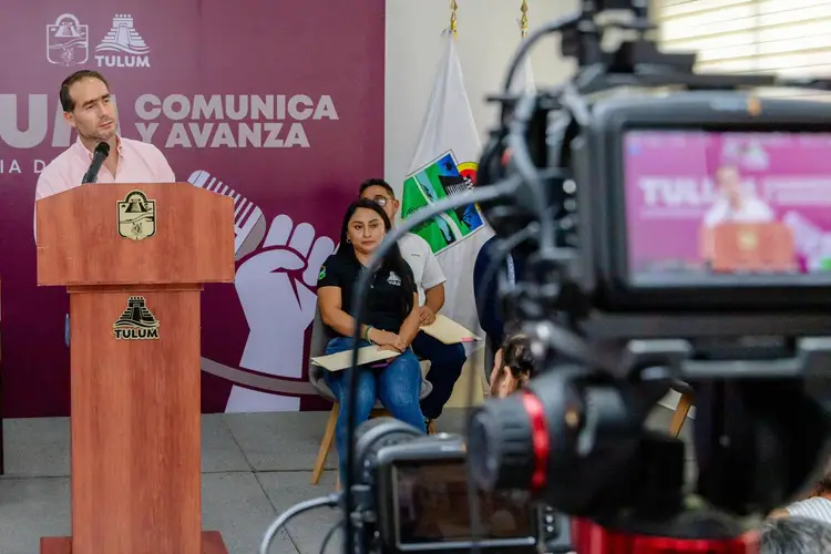 Man speaking at wooden podium with institutional seal while woman sits nearby, camera operator records in foreground, purple banner with text visible behind the