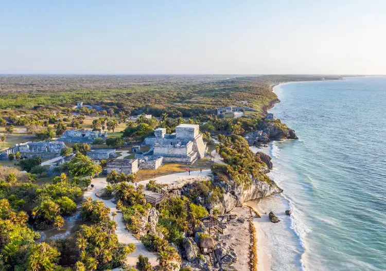 Aerial view of a coastal settlement with white buildings on a clifftop overlooking a beach and turquoise water, surrounded by green vegetation and forest Aerial view of a coastal settlement with white buildings on a clifftop overlooking a beach and turquoise water, surrounded by green vegetation and forest