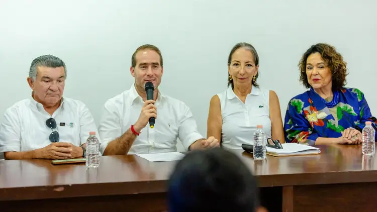 Four panelists sit at a table during what appears to be a public discussion or conference, with the second person from the left speaking into a microphone Four panelists sit at a table during what appears to be a public discussion or conference, with the second person from the left speaking into a microphone