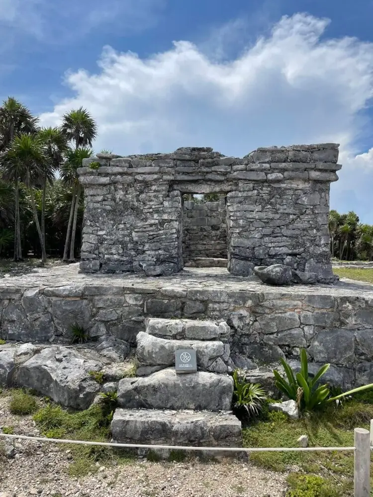 Weathered stone structure with doorway surrounded by palm trees under blue sky with white clouds, with an informational marker visible in the foreground Weathered stone structure with doorway surrounded by palm trees under blue sky with white clouds, with an informational marker visible in the foreground
