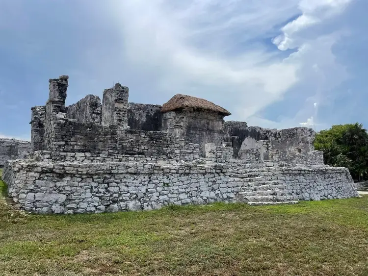 Crumbling stone ruins with a thatched-roof structure stand on a grassy field under a partly cloudy sky Crumbling stone ruins with a thatched-roof structure stand on a grassy field under a partly cloudy sky