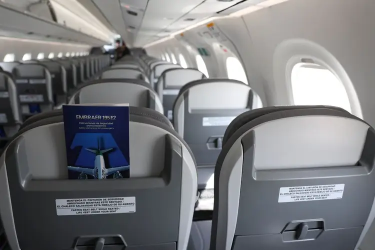 Empty airplane cabin with rows of gray seats, a blue safety card on one seat, and windows along the fuselage Empty airplane cabin with rows of gray seats, a blue safety card on one seat, and windows along the fuselage
