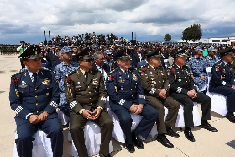 Military and uniformed personnel seated in front rows at an outdoor ceremony with hundreds of attendees gathered behind them on an airfield Military and uniformed personnel seated in front rows at an outdoor ceremony with hundreds of attendees gathered behind them on an airfield
