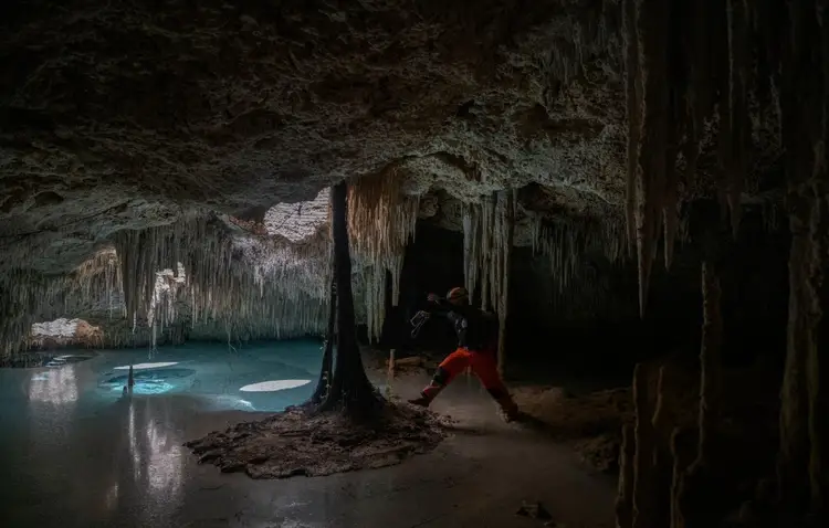 A person in a red shirt stands in a cave chamber with stalactites and a underground pool of blue water A person in a red shirt stands in a cave chamber with stalactites and a underground pool of blue water