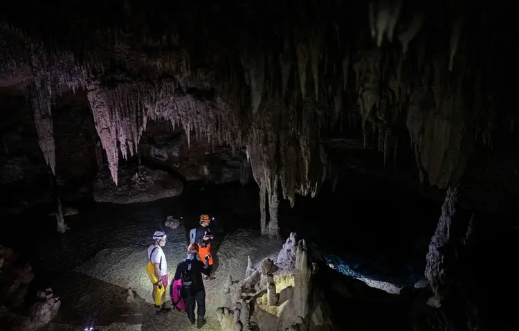 Visitors explore a cave chamber with stalactites hanging from the ceiling and limestone formations on the ground Visitors explore a cave chamber with stalactites hanging from the ceiling and limestone formations on the ground