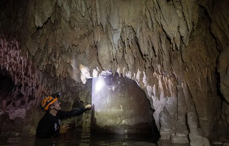 A caver in a yellow helmet shines a light on stalactites and mineral formations inside an underground cave passage filled with water A caver in a yellow helmet shines a light on stalactites and mineral formations inside an underground cave passage filled with water