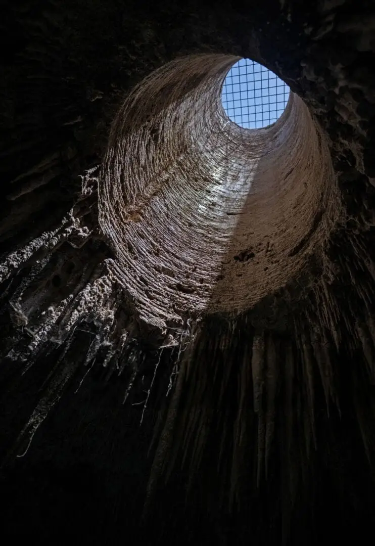 Looking up from inside a deep underground cenote, circular stone walls spiral upward toward a grid-covered opening with blue sky visible above Looking up from inside a deep underground cenote, circular stone walls spiral upward toward a grid-covered opening with blue sky visible above