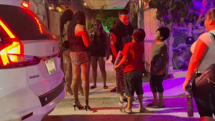 People gather on a street at night illuminated by red and purple neon lights near parked vehicles